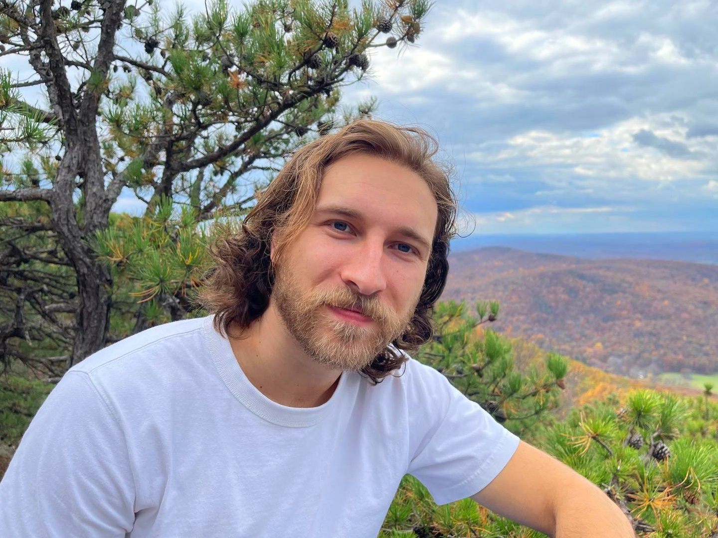 Photo of a dashing gent sitting amongst the brush of a mountain top with rolling hills in the backdrop.